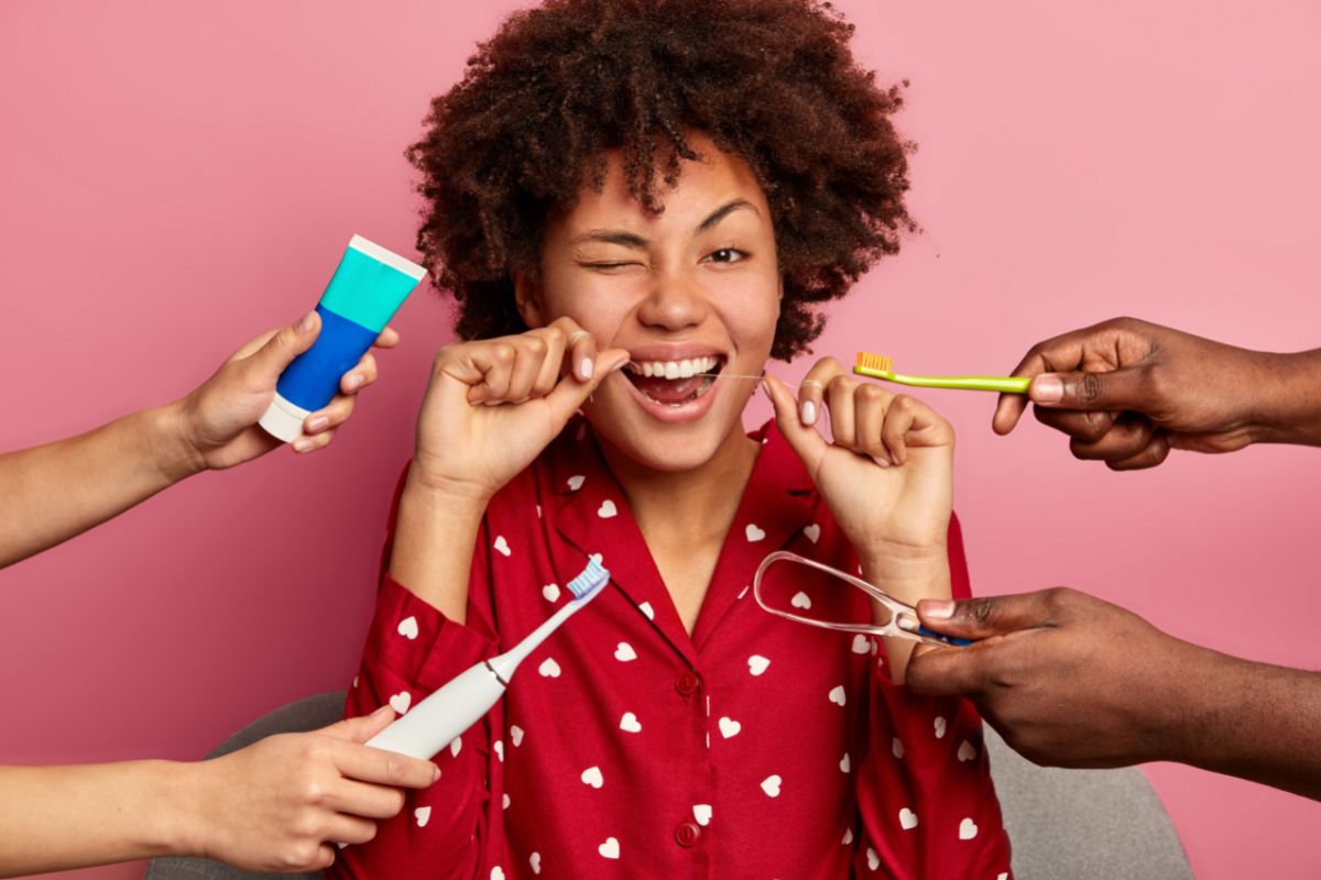 Woman with curly hair smiling while multiple hands offer dental care products against pink background.