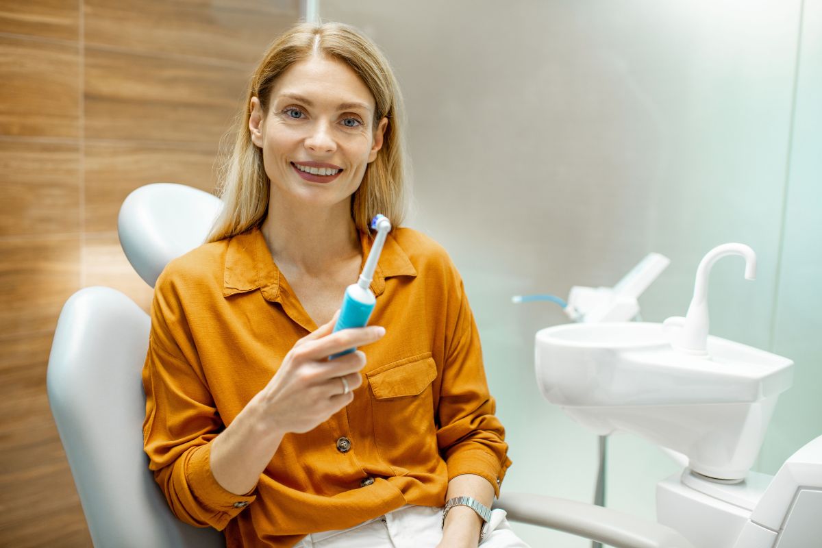 Woman holding toothbrush smiling in dental office with sink visible in background.