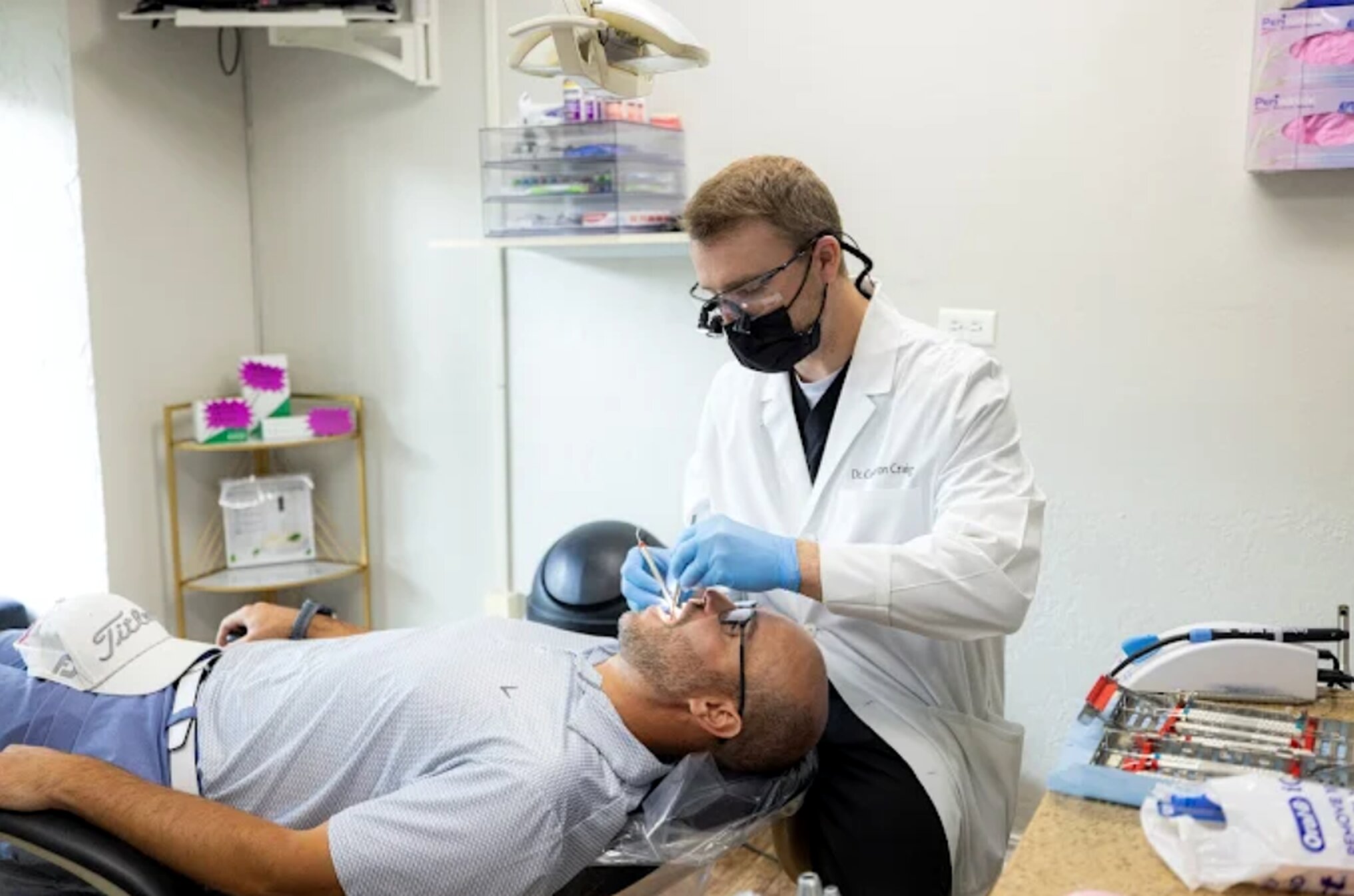 Dentist in white coat and mask examining patient's teeth in modern dental office treatment room.