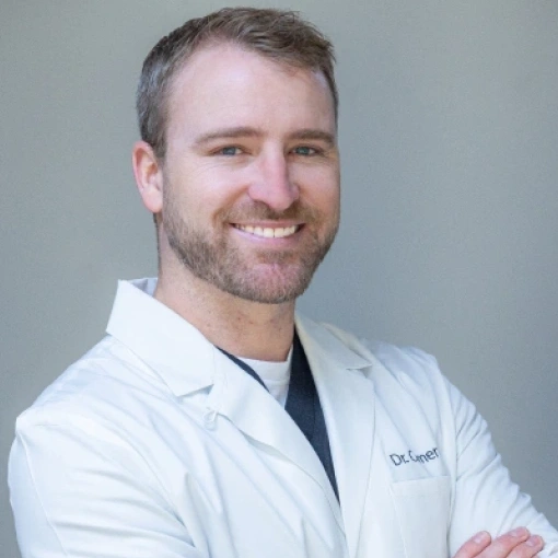 Male dentist in white coat smiling with arms crossed against neutral background.